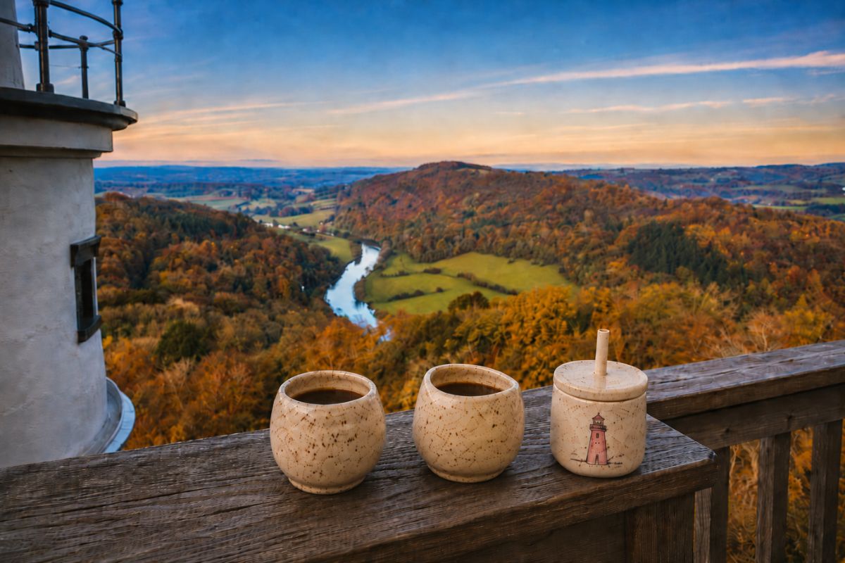 hree speckled ceramic cups on a wooden lighthouse balcony railing overlooking an autumn valley and winding river at sunset, including two coffee mugs and a child’s sippy cup with a pink lighthouse drawing.